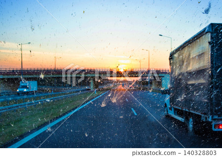 Highway traffic during sunset with trucks and cars through a dirty windshield, showcasing transportation, logistics, road safety and the challenges of visibility while driving on long journeys. Highway traffic during sunset with trucks and cars through a dirty windshield, showcasing transportation, logistics, road safety and the challenges of visibility while driving on long journeys. 140328803