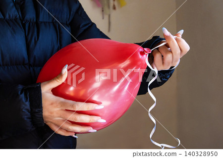 Closeup of woman hands tying ribbon on a red balloon for a party decoration. Person holding a balloon with curly ribbon during birthday party preparations. Closeup of woman hands tying ribbon on a red balloon for a party decoration. Person holding a balloon with curly ribbon during birthday party preparations. 140328950