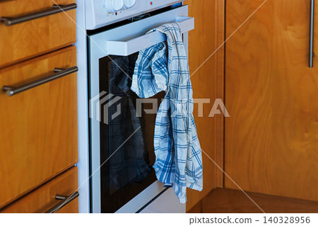 A closeup view of an oven in a domestic kitchen with a crumpled, slightly dirty white blue checkered cotton towel casually hanging from the handle. 140328956