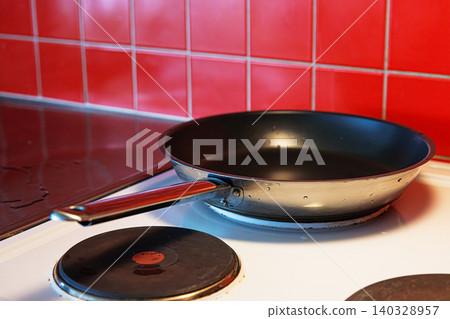 Coseup of a freshly washed clean teflon pan placed on a black cooking plate of a white electric stove with red tile backsplash. Coseup of a freshly washed clean teflon pan placed on a black cooking plate of a white electric stove with red tile backsplash. 140328957
