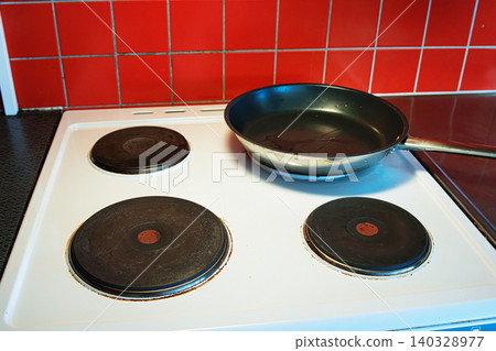 Black nonstick frying pan placed on a white electric stove with four round hotplates in a domestic kitchen with the vibant red tiled backsplash Black nonstick frying pan placed on a white electric stove with four round hotplates in a domestic kitchen with the vibant red tiled backsplash 140328977