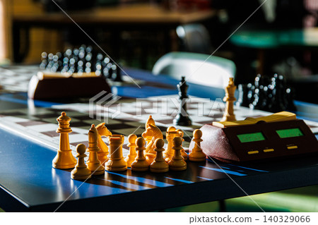 Chess pieces illuminated by sunlight on a tournament table with blurred background. Closeup of a chessboard with a digital timer, showcasing the contrast between light and dark. Selective focus Chess pieces illuminated by sunlight on a tournament table with blurred background. Closeup of a chessboard with a digital timer, showcasing the contrast between light and dark. Selective focus 140329066