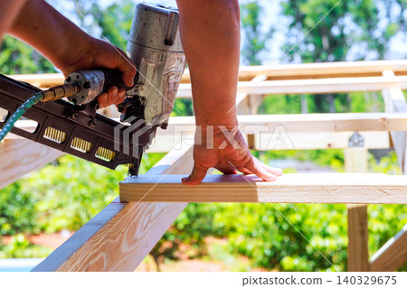 Contractor holds wood steady while using pneumatic nail gun to attach it on construction site 140329675