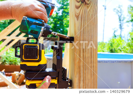 Worker drills screws laser level into wooden post to secure it in place. Worker drills screws laser level into wooden post to secure it in place. 140329679