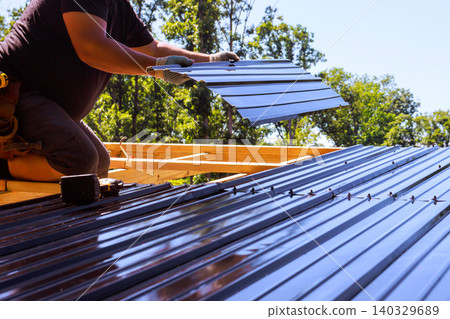 Contractor roofer installs metal roofing panels on wooden beams frame at construction site surrounded by trees. 140329689