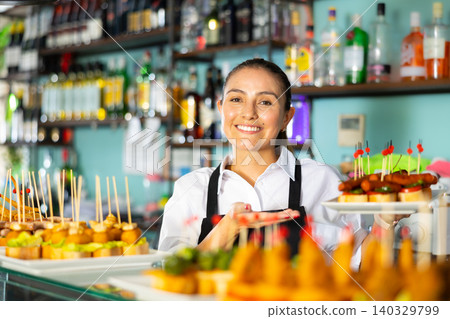 Cheerful Latin female bartender in uniform serving delicious tapas on skewers at the counter of the bar Cheerful Latin female bartender in uniform serving delicious tapas on skewers at the counter of the bar 140329799