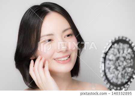 一位年輕女子對著手鏡觸摸自己的皮膚,檢查皮膚狀況,並近距離觀察她的護膚流程。 一位年輕女子對著手鏡觸摸自己的皮膚,檢查皮膚狀況,並近距離觀察她的護膚流程。 140332895