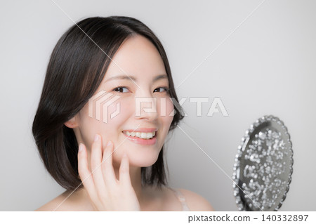 一位年輕女子在進行護膚程序時,對著手鏡檢查自己的肌膚狀況,並對著鏡頭微笑。 一位年輕女子在進行護膚程序時,對著手鏡檢查自己的肌膚狀況,並對著鏡頭微笑。 140332897