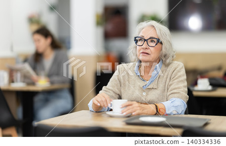 Elderly woman sitting at a table in a coffee shop with a cup of aromatic coffee Elderly woman sitting at a table in a coffee shop with a cup of aromatic coffee 140334336