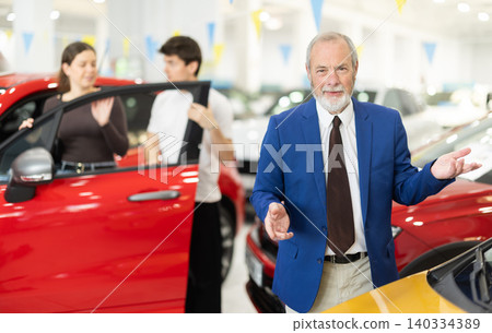 Elderly salesman stands in a car dealership and invites customers to see the cars Elderly salesman stands in a car dealership and invites customers to see the cars 140334389