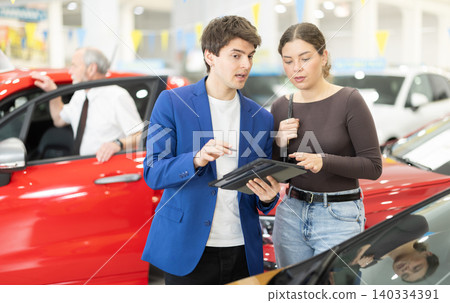 Young auto salesman advises a woman, answering her questions and showing detailed information about the car Young auto salesman advises a woman, answering her questions and showing detailed information about the car 140334391