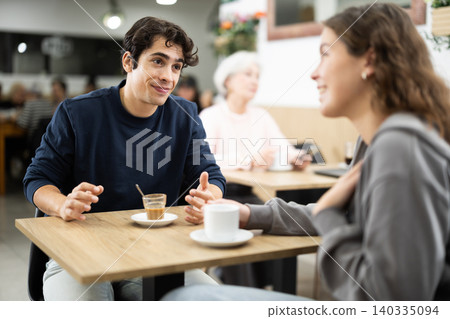 Happy young guy and pretty girl on date over cup of coffee at table in light cafe, face of guy and back of girl Happy young guy and pretty girl on date over cup of coffee at table in light cafe, face of guy and back of girl 140335094