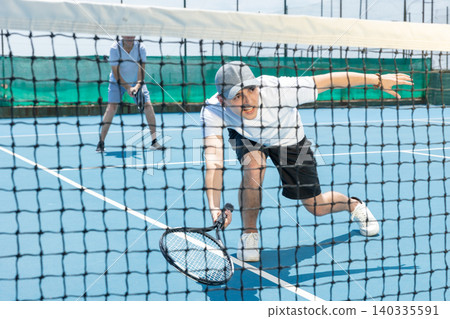 Portrait of young guy playing tennis on open court in summer, swinging racket to return ball over net. Sportsman ready to hit volley Portrait of young guy playing tennis on open court in summer, swinging racket to return ball over net. Sportsman ready to hit volley 140335591