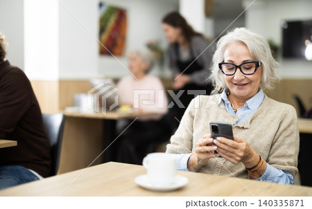European elderly woman drinking fresh coffee at table in cafe and looking at mobile phone 140335871