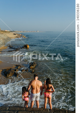 Rear View of Father and Two Daughters Standing by Ocean Waves on Rocky Beach at Sunset Rear View of Father and Two Daughters Standing by Ocean Waves on Rocky Beach at Sunset 140336180