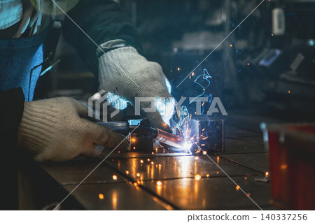 A craftsman's hands work as he welds at a workbench in a small factory, illuminated by a blue light and amidst the intensely flying sparks. A craftsman's hands work as he welds at a workbench in a small factory, illuminated by a blue light and amidst the intensely flying sparks. 140337256