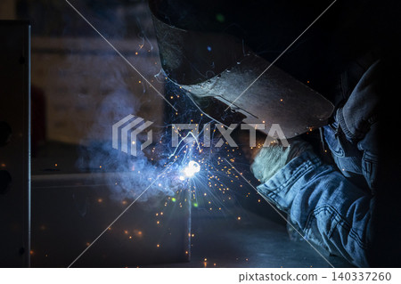 A worker wearing a welding mask performs welding in the dark, creating intense sparks and smoke. (Close-up) 140337260