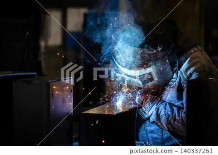 一名戴著焊接面罩的焊工在一家小型工廠昏暗的車間工作，藍色的煙霧和火花四處飛濺。 140337261
