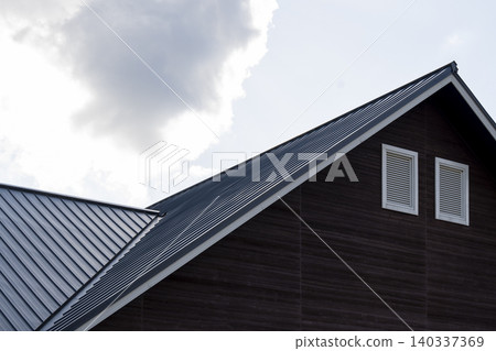 A modern building facade with a metal corrugated roof and brown wood-grain exterior walls against a cloudy sky. A modern building facade with a metal corrugated roof and brown wood-grain exterior walls against a cloudy sky. 140337369