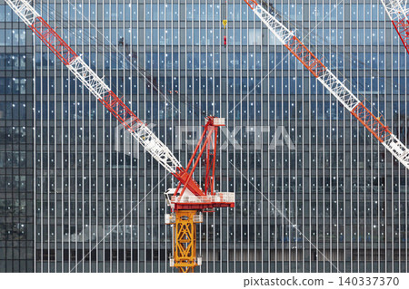 在現代建築的玻璃帷幕牆背景下，建築工地上的大型塔式起重機巍然聳立。 140337370