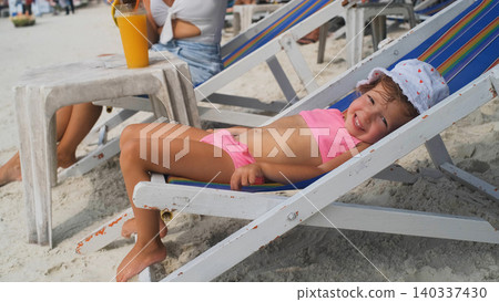 Cute little girl in pink swimsuit lying and relaxing on a beach lounge chair on a sunny tropical beach. Childhood vacation concept Cute little girl in pink swimsuit lying and relaxing on a beach lounge chair on a sunny tropical beach. Childhood vacation concept 140337430