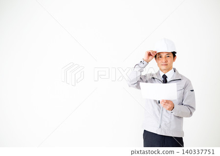 A middle-aged businessman in gray work clothes and a helmet is looking at documents against a white background. A middle-aged businessman in gray work clothes and a helmet is looking at documents against a white background. 140337751