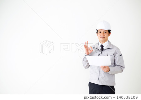 A middle-aged businessman in gray work clothes and a helmet, looking at a document and making a circle sign against a white background. A middle-aged businessman in gray work clothes and a helmet, looking at a document and making a circle sign against a white background. 140338109