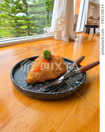 Delicious baked croissant topped with creamy mentaiko sauce and ebiko flying fish roe served on a black ceramic plate on a wooden table in a cafe. Delicious baked croissant topped with creamy mentaiko sauce and ebiko flying fish roe served on a black ceramic plate on a wooden table in a cafe. 140338614