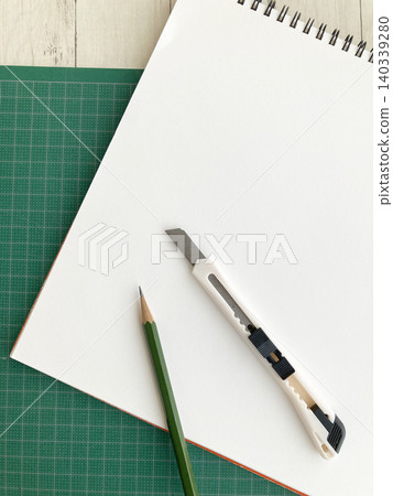 A scene of crafting and design work; a sketchbook, utility knife, and pencil on a cutting mat; office supplies. A scene of crafting and design work; a sketchbook, utility knife, and pencil on a cutting mat; office supplies. 140339280