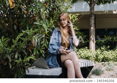 Woman sits on outdoor bench talking on mobile phone next to laptop computer and holding notebook. Mobile business communication, client relations call, remote consulting conversation, phone interview Woman sits on outdoor bench talking on mobile phone next to laptop computer and holding notebook. Mobile business communication, client relations call, remote consulting conversation, phone interview 140340208