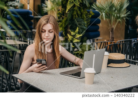 Young woman sits at outdoor cafe table using smartphone and laptop. Cross platform communication, synchronized digital tasks, multitasking female professional. Young woman sits at outdoor cafe table using smartphone and laptop. Cross platform communication, synchronized digital tasks, multitasking female professional. 140340222