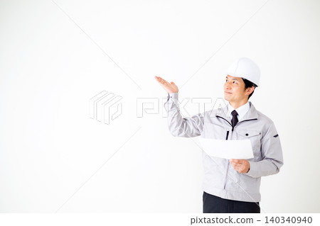 A middle-aged businessman in gray work clothes and a helmet is looking at documents against a white background. A middle-aged businessman in gray work clothes and a helmet is looking at documents against a white background. 140340940