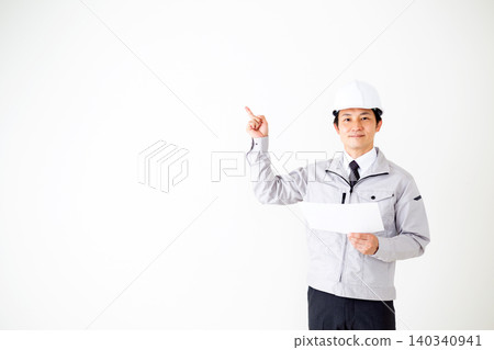 A middle-aged businessman in gray work clothes and a helmet is looking at documents against a white background. A middle-aged businessman in gray work clothes and a helmet is looking at documents against a white background. 140340941