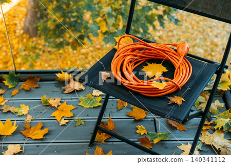 chair stands on wooden balcony surrounded by autumn leaves with coiled orange extension cord on seat. Autumn chores routine, DIY home maintenance, Preparing house for winter, outdoor electrical setup chair stands on wooden balcony surrounded by autumn leaves with coiled orange extension cord on seat. Autumn chores routine, DIY home maintenance, Preparing house for winter, outdoor electrical setup 140341112