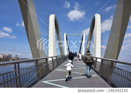 Adults and children walk and ride scooters across a modern arched pedestrian bridge by the river. A family outing on a sunny spring day on a contemporary city bridge. Adults and children walk and ride scooters across a modern arched pedestrian bridge by the river. A family outing on a sunny spring day on a contemporary city bridge. 140342235