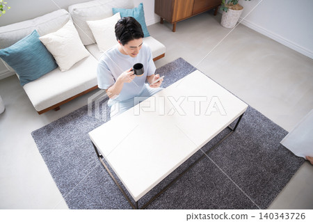 A bird's-eye view of a man's lifestyle: drinking coffee in the living room while using his smartphone. 140343726
