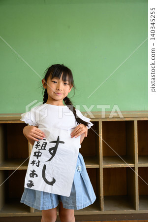 A photo of a female elementary school student smiling and showing her calligraphy artwork of the word "sushi" at the back of the classroom. A photo of a female elementary school student smiling and showing her calligraphy artwork of the word "sushi" at the back of the classroom. 140345255