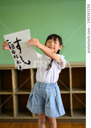 一張照片中,一名小學生面帶微笑,在教室後方展示她用書法寫成的「壽司」字。 一張照片中,一名小學生面帶微笑,在教室後方展示她用書法寫成的「壽司」字。 140345256