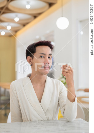 Vertical portrait of non-binary gay Thai man looking at camera sitting at a modern cafe table Vertical portrait of non-binary gay Thai man looking at camera sitting at a modern cafe table 140345397