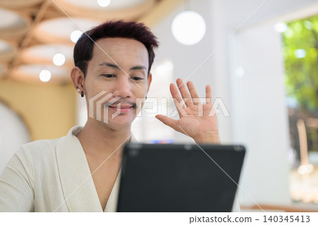 Close up portrait of non-binary gay Thai man waving at digital tablet screen during cafe video call Close up portrait of non-binary gay Thai man waving at digital tablet screen during cafe video call 140345413