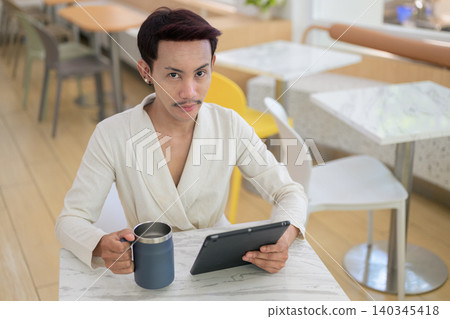 High angle portrait of non-binary gay Thai man holding coffee mug and digital tablet at cafe table 140345418
