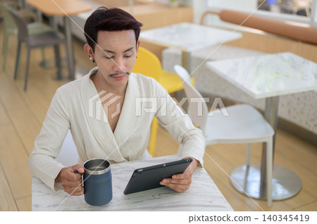 High angle view of non-binary gay Thai man looking down at tablet screen holding coffee mug High angle view of non-binary gay Thai man looking down at tablet screen holding coffee mug 140345419