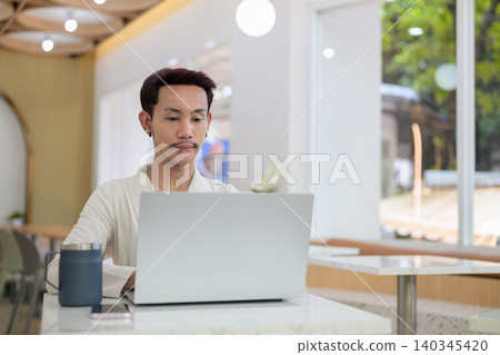 Focused non-binary gay Thai man typing on laptop while working remotely at modern cafe table 140345420