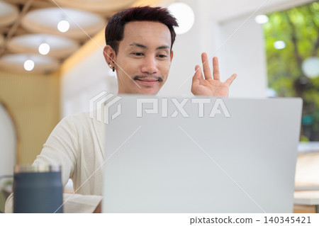 Close up portrait of smiling non-binary gay Thai man waving at laptop screen during cafe video call Close up portrait of smiling non-binary gay Thai man waving at laptop screen during cafe video call 140345421