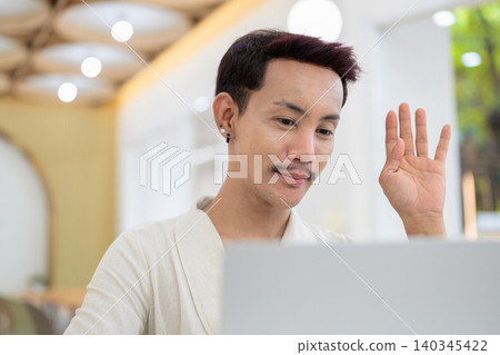 Close up portrait of non-binary gay Thai man waving hand during video call on laptop at cafe Close up portrait of non-binary gay Thai man waving hand during video call on laptop at cafe 140345422