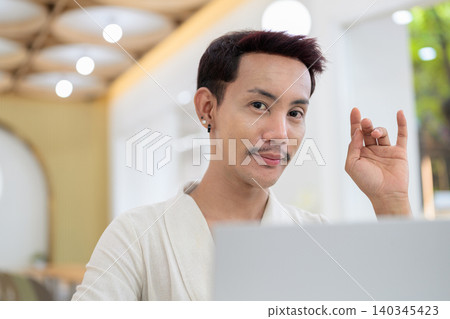 Portrait of smiling non-binary gay Thai man making I love you hand gesture during laptop video call 140345423