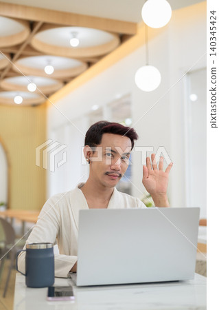 Vertical portrait of non-binary gay Thai man waving hand while looking at camera behind laptop at cafe Vertical portrait of non-binary gay Thai man waving hand while looking at camera behind laptop at cafe 140345424