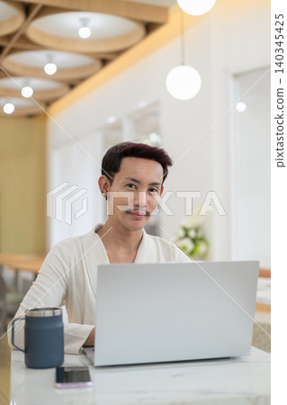 Vertical portrait of non-binary gay Thai man smiling at camera while working on laptop at cafe Vertical portrait of non-binary gay Thai man smiling at camera while working on laptop at cafe 140345425