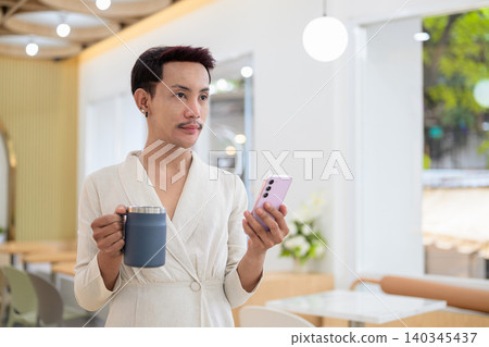 Non-binary gay Thai man looking away while holding smartphone and coffee mug in modern cafe Non-binary gay Thai man looking away while holding smartphone and coffee mug in modern cafe 140345437