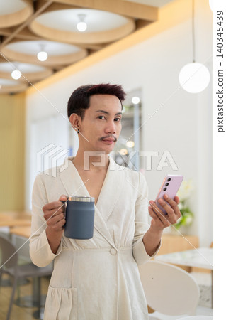 Vertical portrait of non-binary gay Thai man looking at camera while holding smartphone and coffee mug Vertical portrait of non-binary gay Thai man looking at camera while holding smartphone and coffee mug 140345439